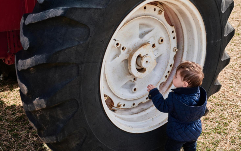 Un niño vestido de azul junto a un neumático de tractor grande, con curiosidad